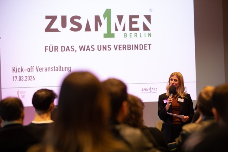 Frau spricht vor einem Publikum, hinter ihr ein Bildschirm mit dem Zusammen1 Logo.