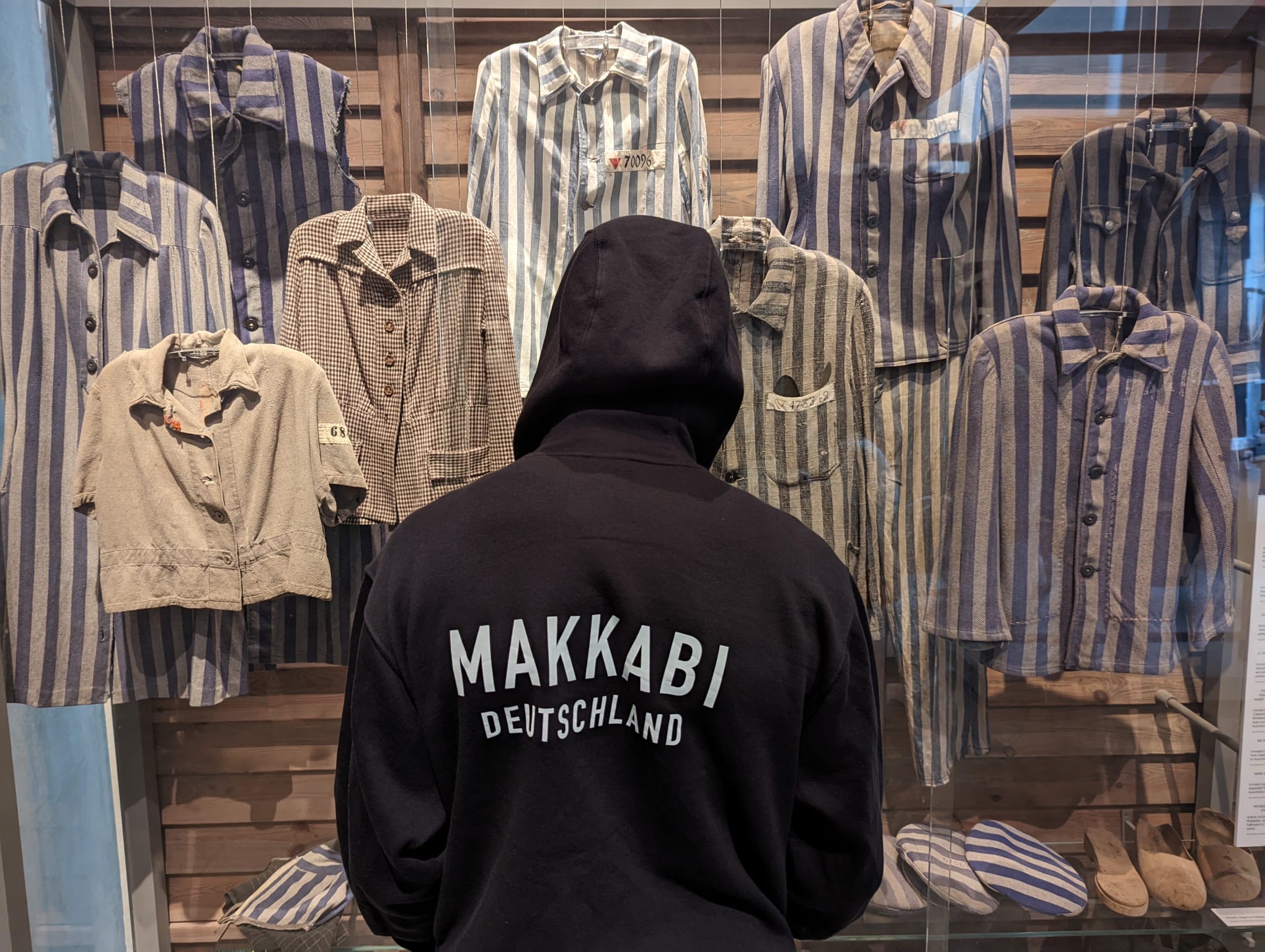 Person in schwarzem Hoodie mit der Aufschrift „Makkabi Deutschland“ betrachtet eine Ausstellung historischer gestreifter Häftlingskleidung hinter Glas.