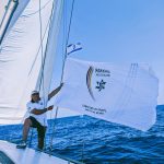 Ausschnitt eines Segelbootes auf dem Meer, ein Sportler hält die wehende Makkabi Deutschland Flagge